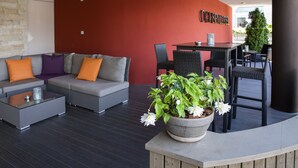 Un espace de détente extérieur moderne, avec un canapé gris, des coussins orange, une table basse en verre et une plante en pot ornée de fleurs blanches.
