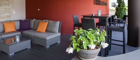 Ein moderner Außenbereich mit einer grauen Couch, orangefarbenen Kissen, einem Glastisch und einem Blumentopf mit weißen Blüten.