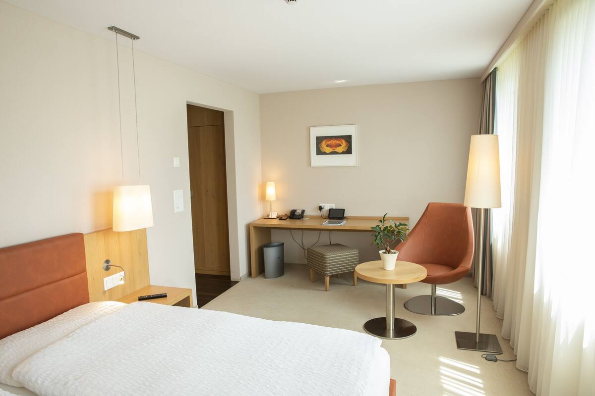 A hotel room with a bed, desk, chair, and a potted plant.