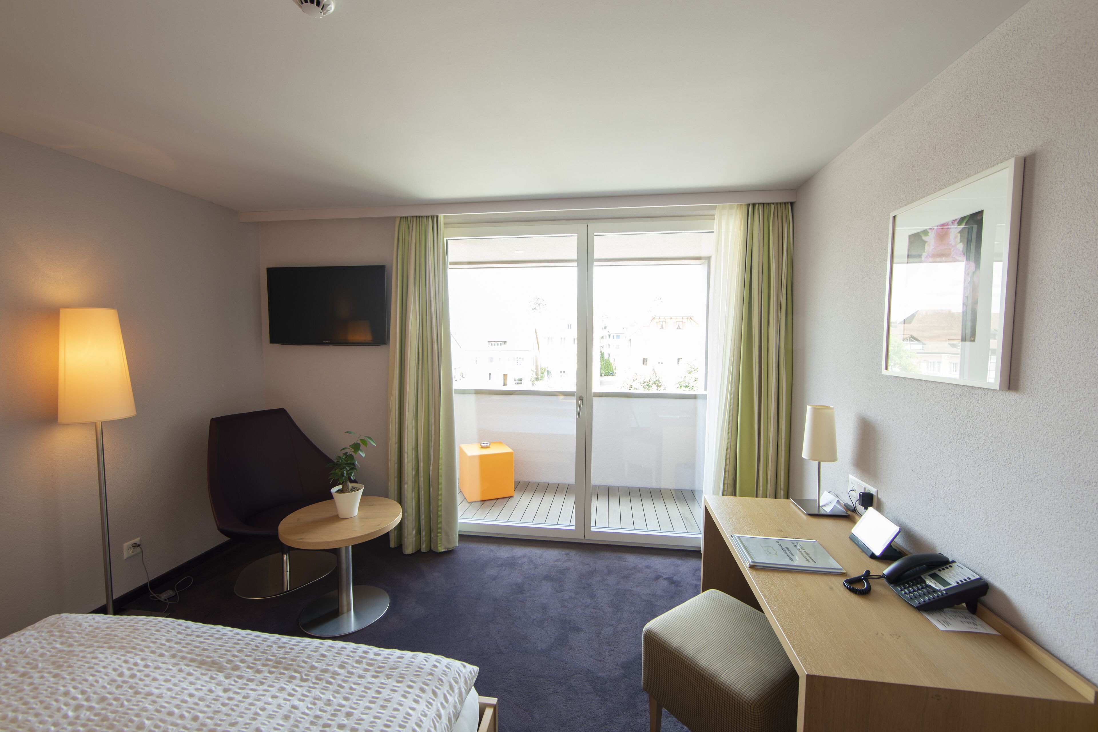 Et hotelværelse med en seng, et skrivebord, en stol, et lille bord, en plante, et fjernsyn og en balkon med gardiner.