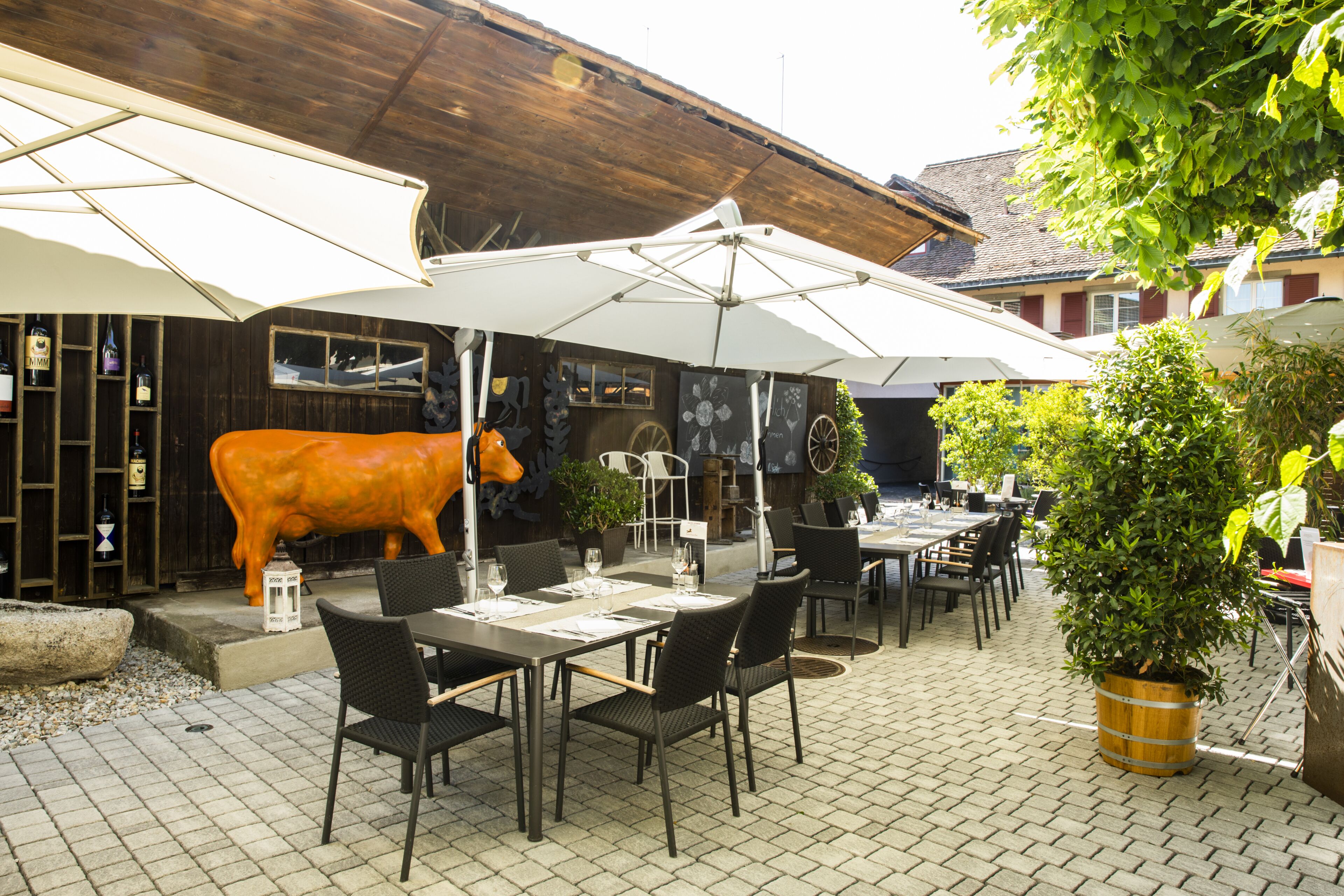 Ein Außenbereich zum Essen mit Tischen, Stühlen und Sonnenschirmen, sowie eine große Kuhstatue.