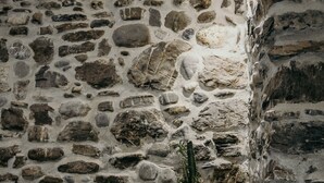 Un muro de piedra con un cactus en una maceta blanca, una silla azul y una ventana pequeña.