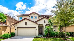 A two-story house with a beige facade, a brown roof, and a garage. There are trees and shrubs in the front yard. - Stunning villa, lake views-south facing pool - game room,  near Disney & golf (Davenport)