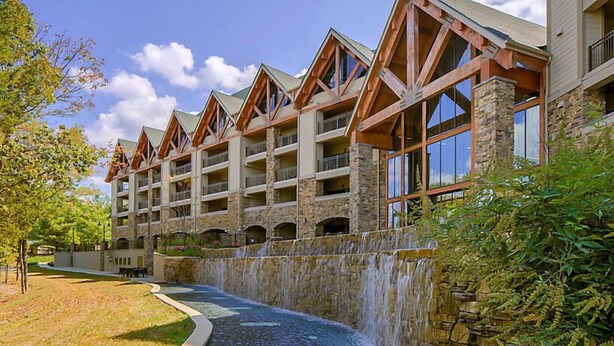 Um hotel com vários andares, fachada em pedra e madeira, um recurso com cascata e um pátio ajardinado.