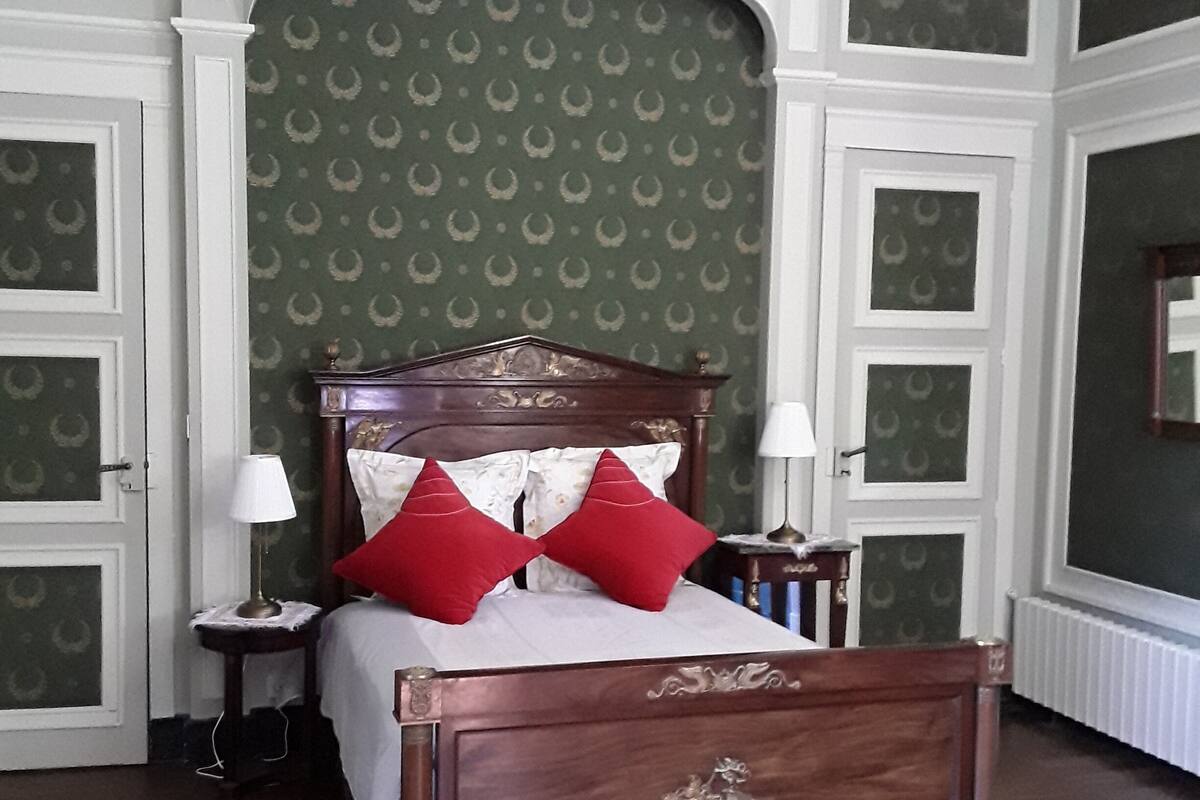 Un lit à baldaquin classique, agrémenté d’oreillers rouges, d’une tête de lit en bois et d’un papier peint à motifs verts.