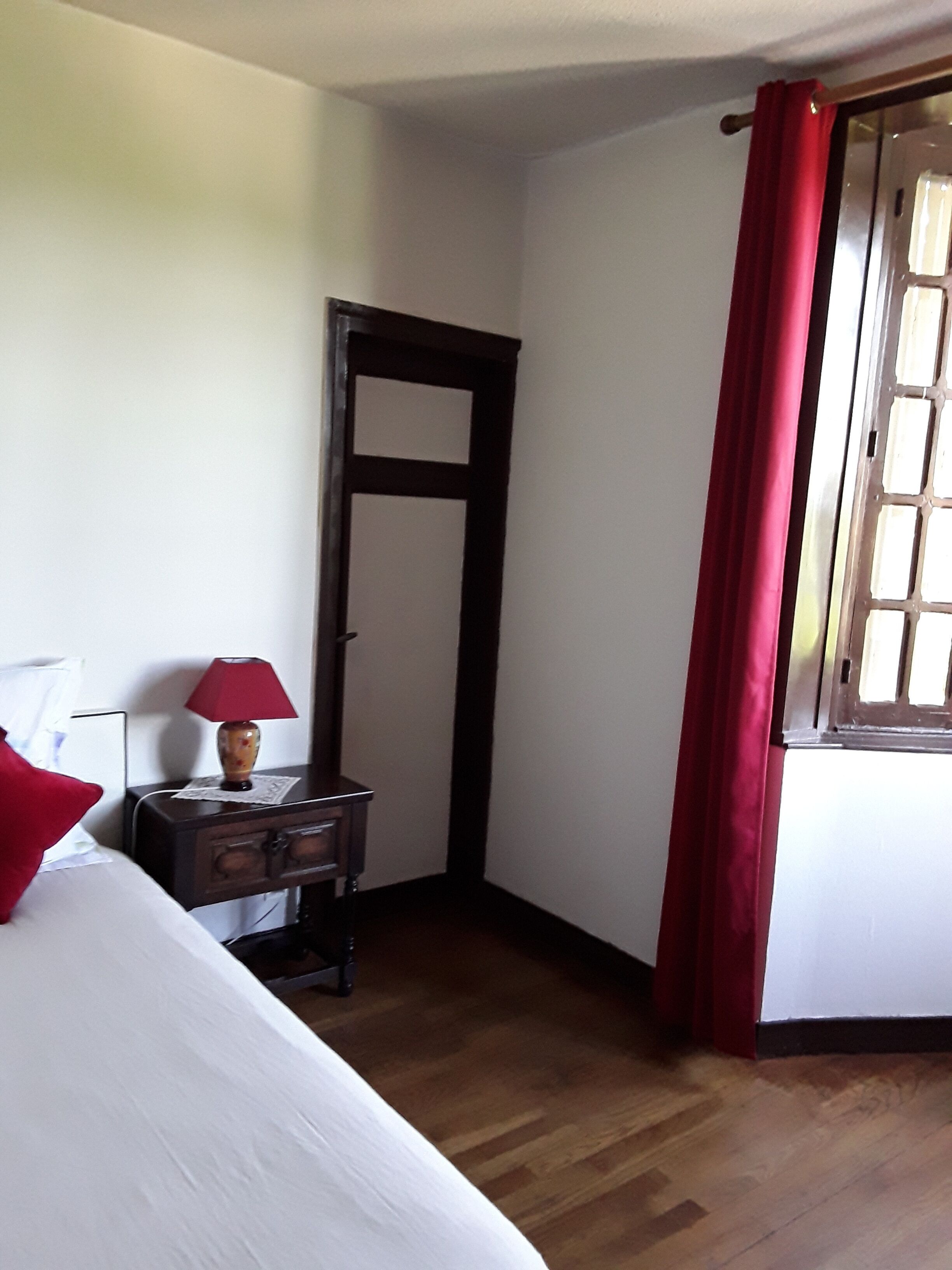 Une chambre d’hôtel avec un lit, une table de chevet avec une lampe, une porte en bois et une fenêtre avec des rideaux rouges.