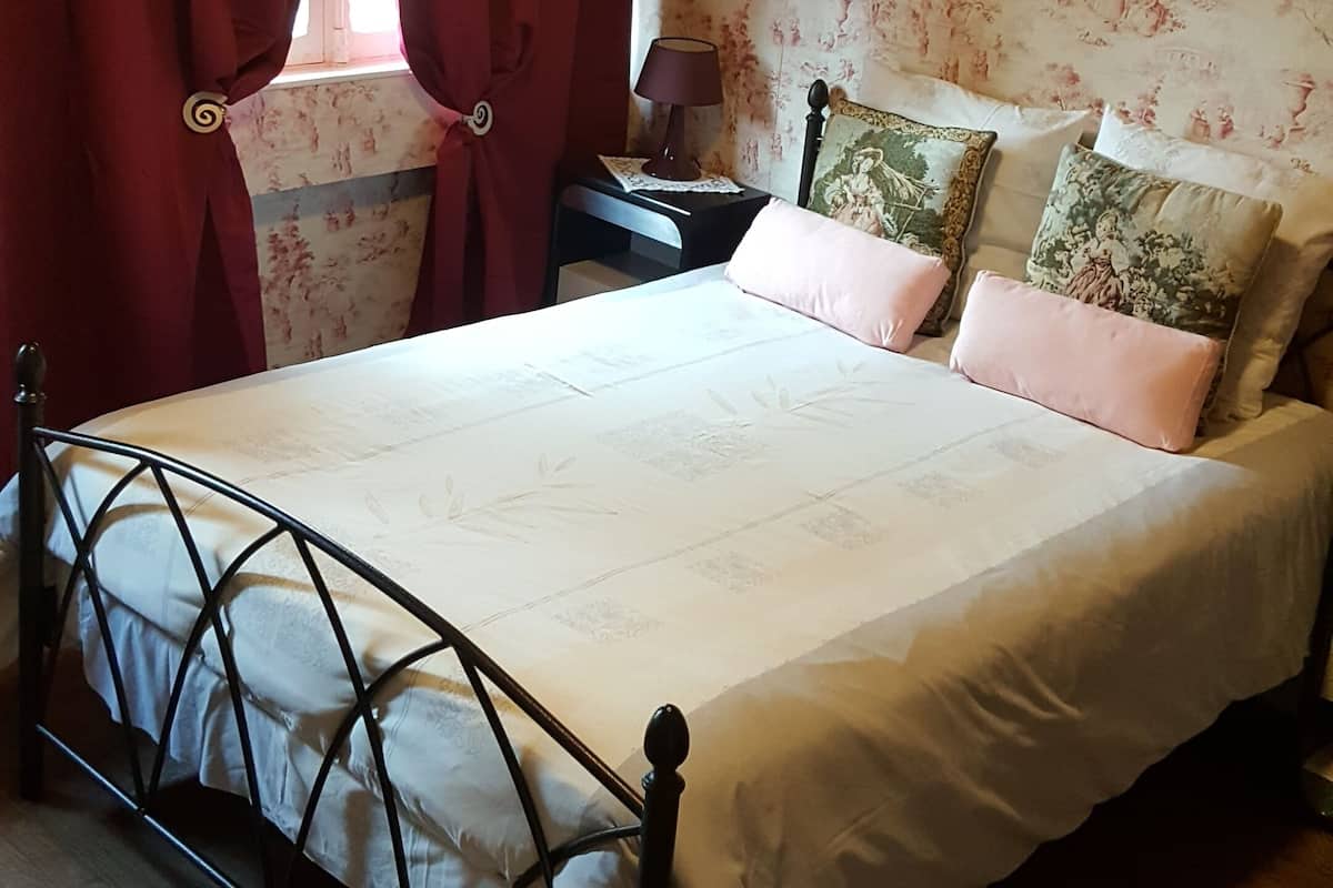 Une chambre à coucher avec un lit, des rideaux, une table de chevet, une lampe et un mur recouvert d’un papier peint à motifs floraux.