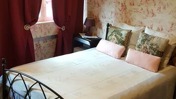 Un dormitorio con cama, cortinas, una mesita de noche, lámpara y un papel tapiz floral en la pared.
