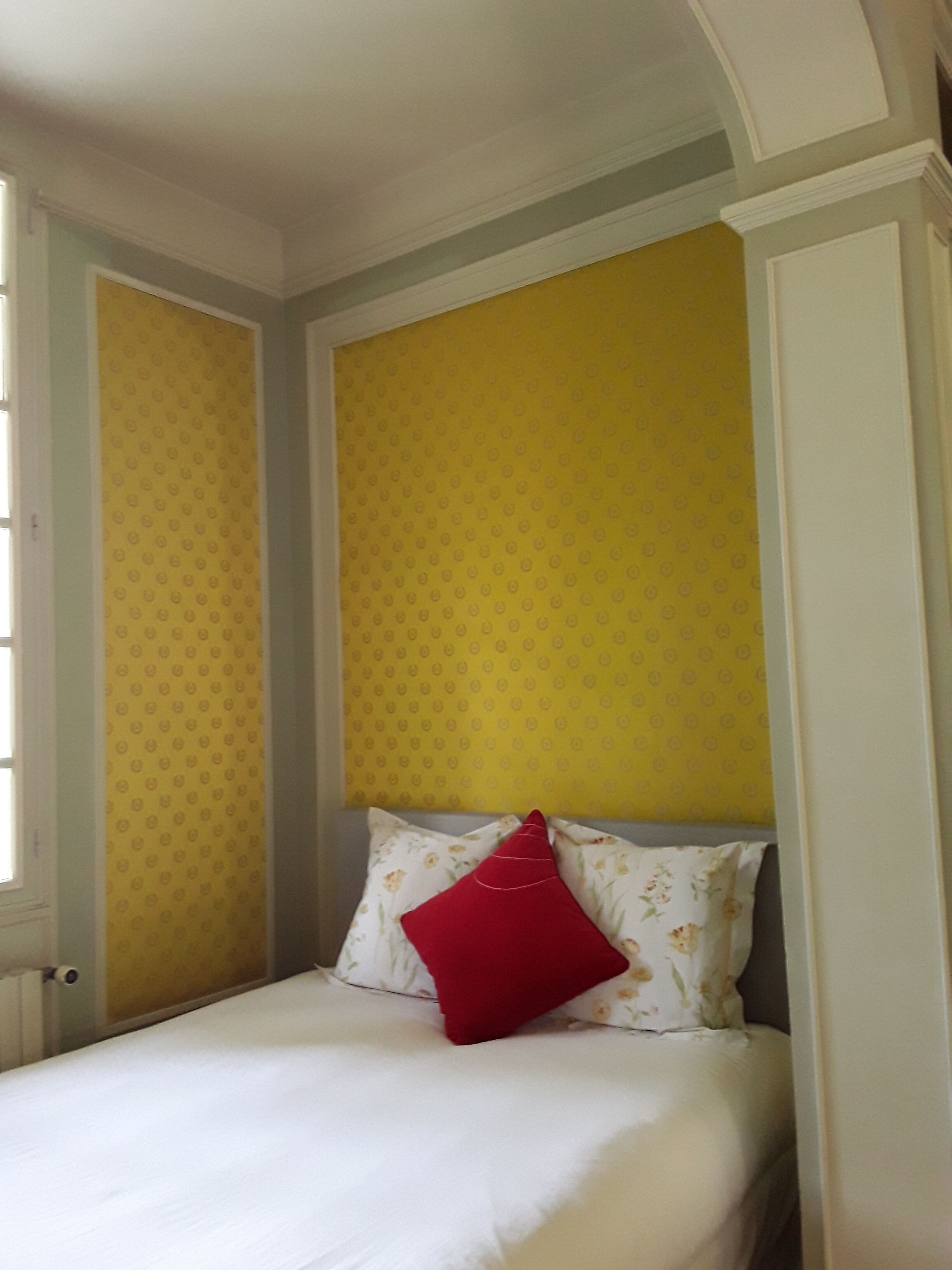 A single bed with a white mattress and a red pillow, a yellow patterned wall, and a white door.
