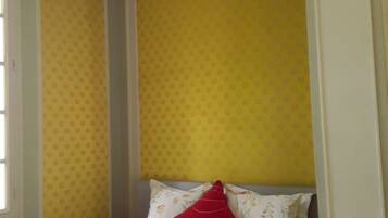A single bed with a white mattress and a red pillow, a yellow patterned wall, and a white door.