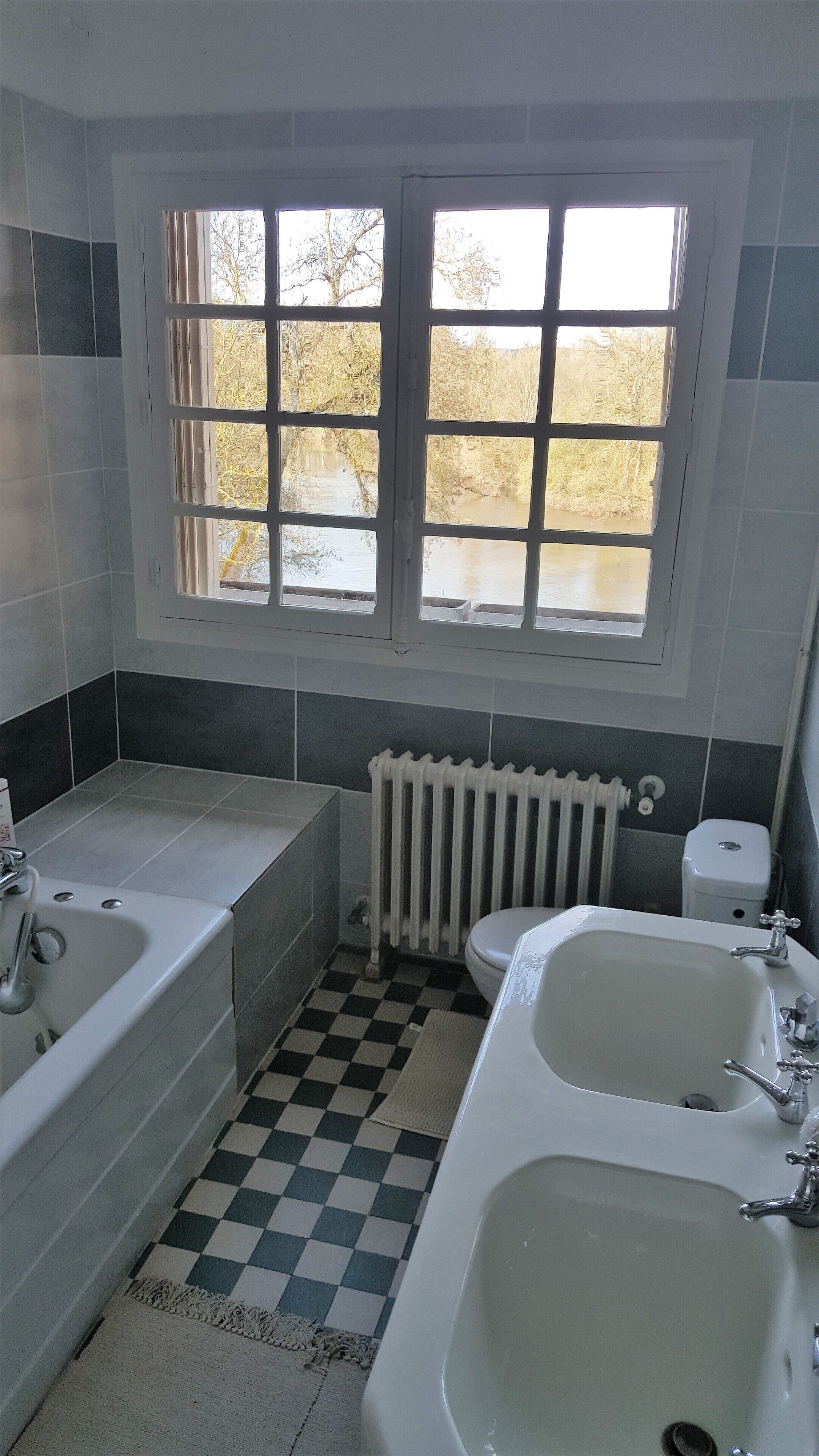 A bathroom with a checkered floor, two sinks, a bathtub, a toilet, and a window with a view of trees.