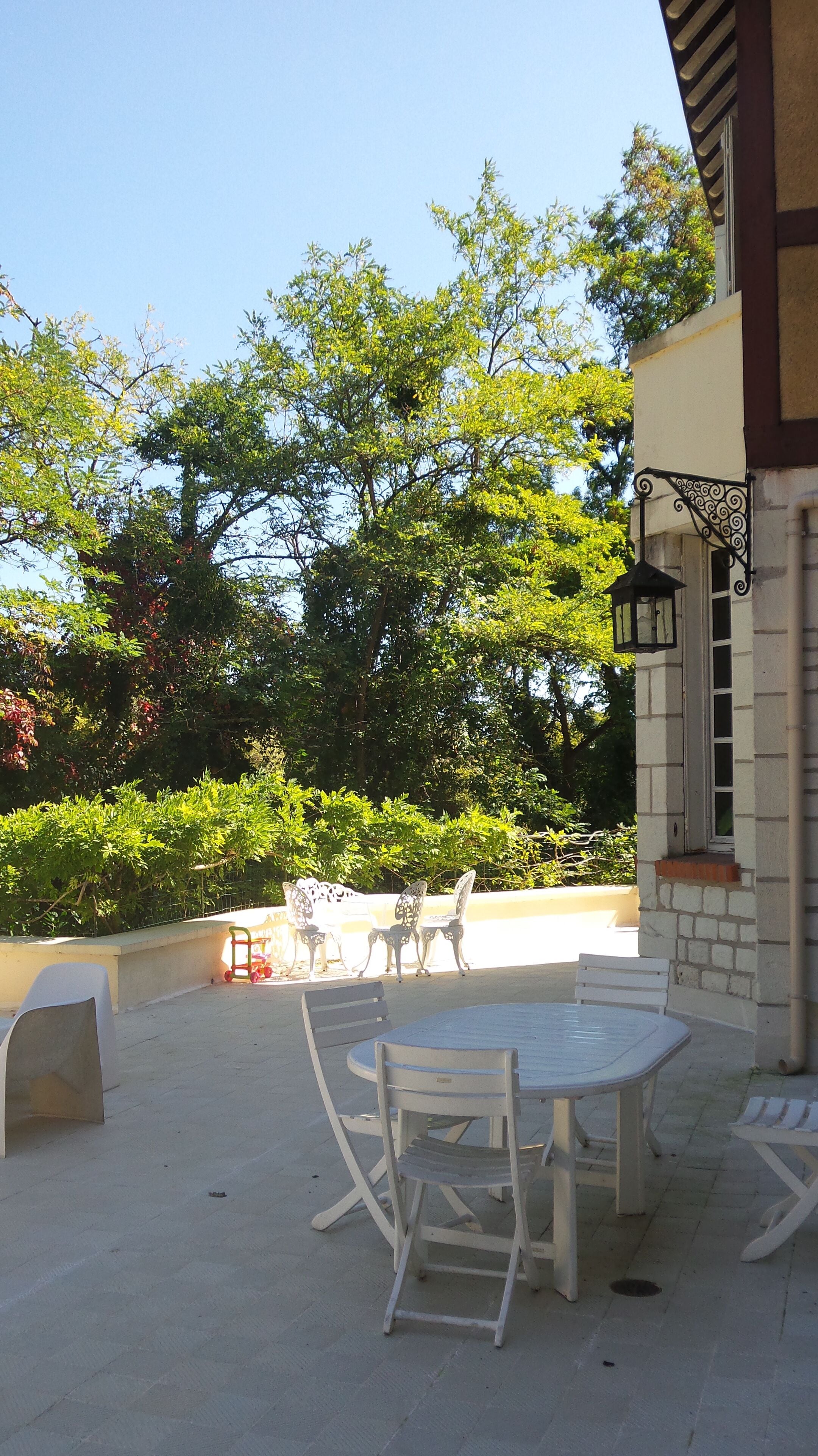 An outdoor patio with a round table and white chairs, surrounded by greenery and trees.