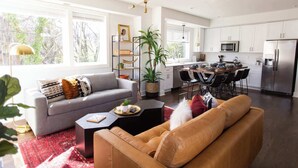 Una sala moderna con un sofá gris, un sofá de cuero marrón y una mesa de centro. Hay una zona de cocina con armarios blancos, un refrigerador de acero inoxidable y una mesa de comedor con sillas.
