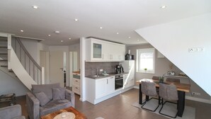 A modern kitchen with white cabinets, a dining area with a wooden table and chairs, a gray sofa, and a staircase leading to another level. - Apartment Hafenbrise in Greetsiel for 4 people (Greetsiel)