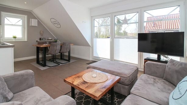 A modern living room with a sofa, a coffee table, and a TV. There is a dining area with a table and chairs. - Apartment Hafenbrise in Greetsiel for 4 people (Greetsiel)