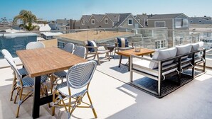 A rooftop patio with a wooden table and wicker chairs, a sofa with cushions, and a view of residential buildings. - Book now Summer is here— Pet,2BR/2BA, 2 Parking (Newport Beach)