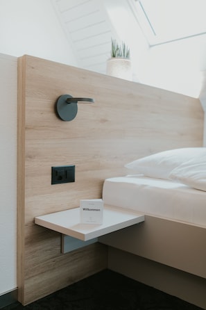 A modern bedroom with a wooden headboard, a bedside table with a welcome card, and a small potted plant on the wall.