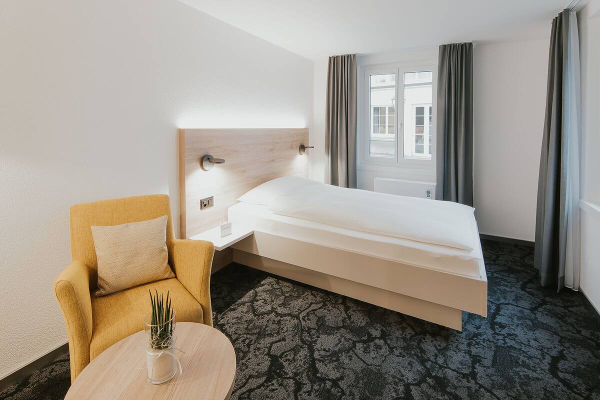 Ein Hotelzimmer mit einem Bett, einem Sessel, einem kleinen Tisch und einem Fenster mit Vorhängen.