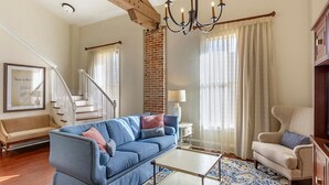 Un salon avec un canapé bleu, un fauteuil beige et une table basse. On aperçoit un escalier avec une rampe en bois à l’arrière-plan.
