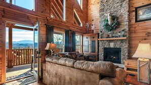 A cozy cabin interior with a stone fireplace, leather sofa, and a deck with a view of the mountains. - WHEELCHAIR ACCESSIBLE, 4 King Suites, Close to Pigeon Forge, WOW Views! (Sevierville)