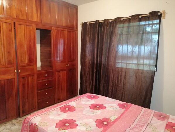 A bedroom with a wooden wardrobe, a bed with a floral bedspread, and a window with a curtain.