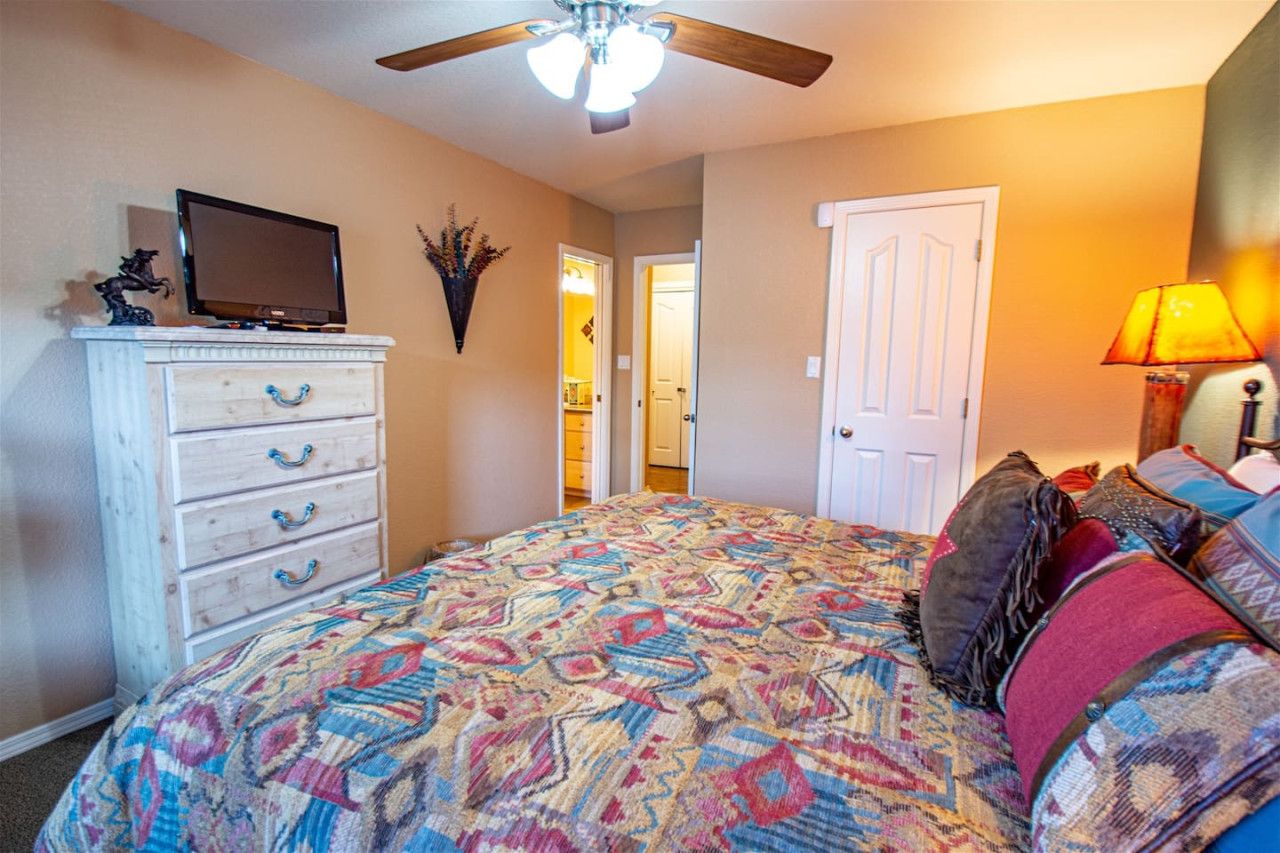 A bedroom with a patterned bedspread, a television on a dresser, a ceiling fan, and a door leading to another room.