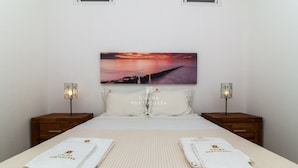 A hotel room with a bed, two nightstands, a painting of a sunset, and towels with "D'ALMA PORTUGUESA" text.