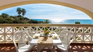 A table set for two with white chairs, a vase with flowers, and plates with food, overlooking a coastal view with palm trees and the sea.