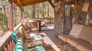 A covered outdoor seating area with a stone wall, a wooden deck, and a view of the surrounding forest. - Horsin` Around - Unit M2 (Ruidoso)