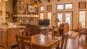 A spacious living area with a stone fireplace, wooden furniture, and large windows. - Horsin` Around - Unit M2 (Ruidoso)