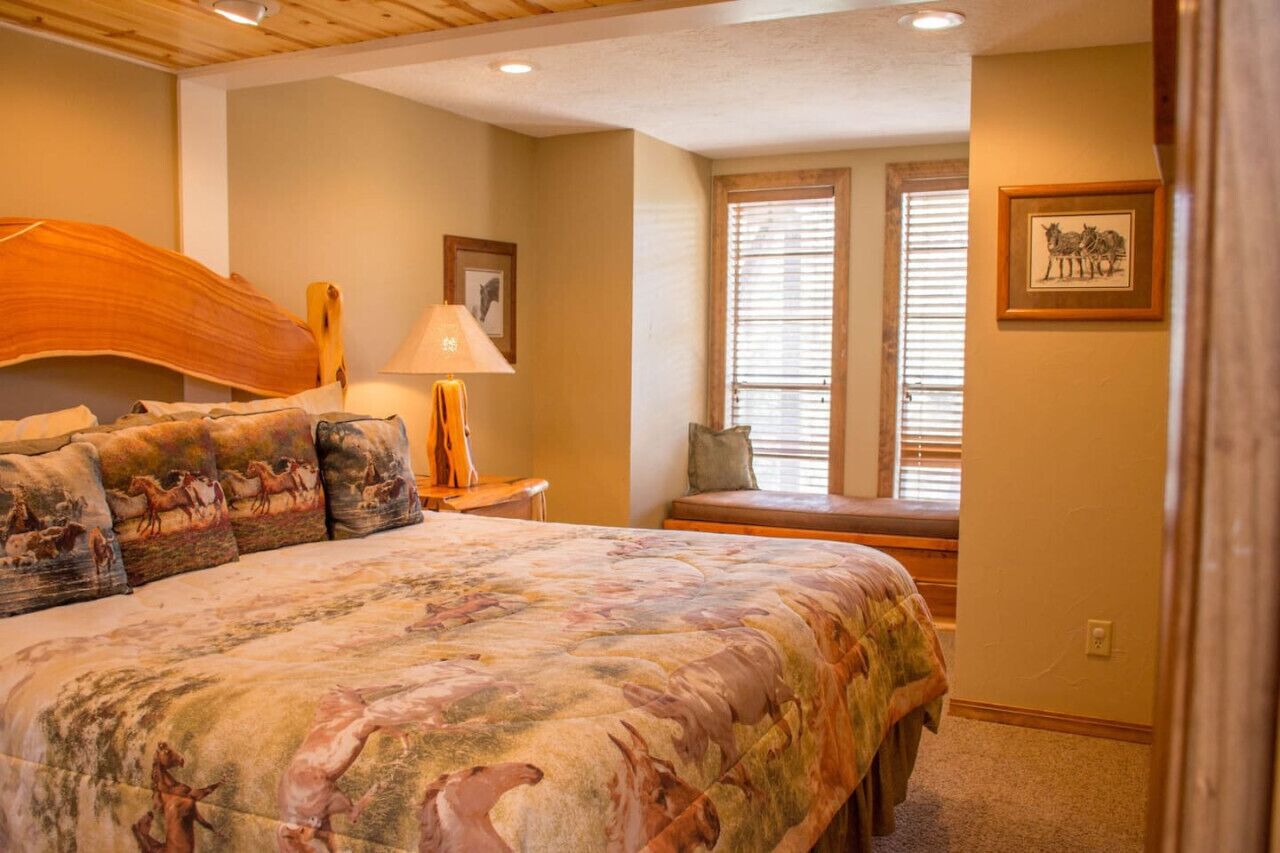 A bedroom with a large bed, a nightstand, a lamp, a window with blinds, and a framed picture on the wall.