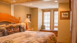 A bedroom with a large bed, a nightstand, a lamp, a window with blinds, and a framed picture on the wall. - Horsin` Around - Unit M2 (Ruidoso)