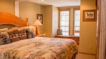 A bedroom with a large bed, a nightstand, a lamp, a window with blinds, and a framed picture on the wall.