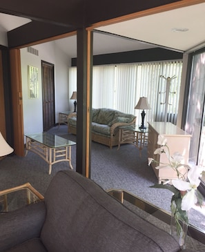 A living room with a sofa, a glass coffee table, a wicker chair, and a lamp. There is a view of a bedroom with a bed and curtains.