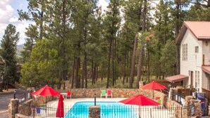 A swimming pool surrounded by stone walls, with red umbrellas and lounge chairs. - Come Stay in Antlers - D4 (Ruidoso)