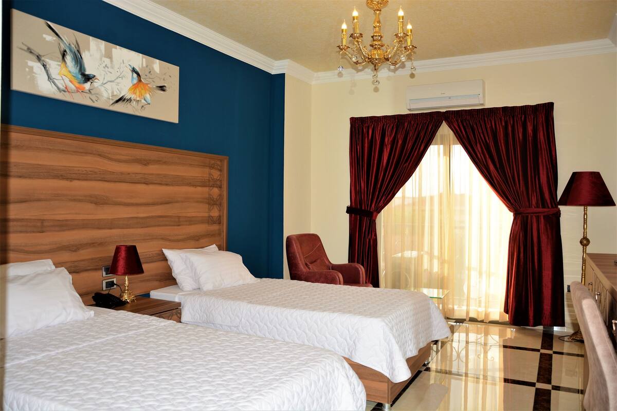 Een hotelkamer met twee bedden, een houten hoofdbord, een kroonluchter, een rode stoel en een groot raam met rode gordijnen.