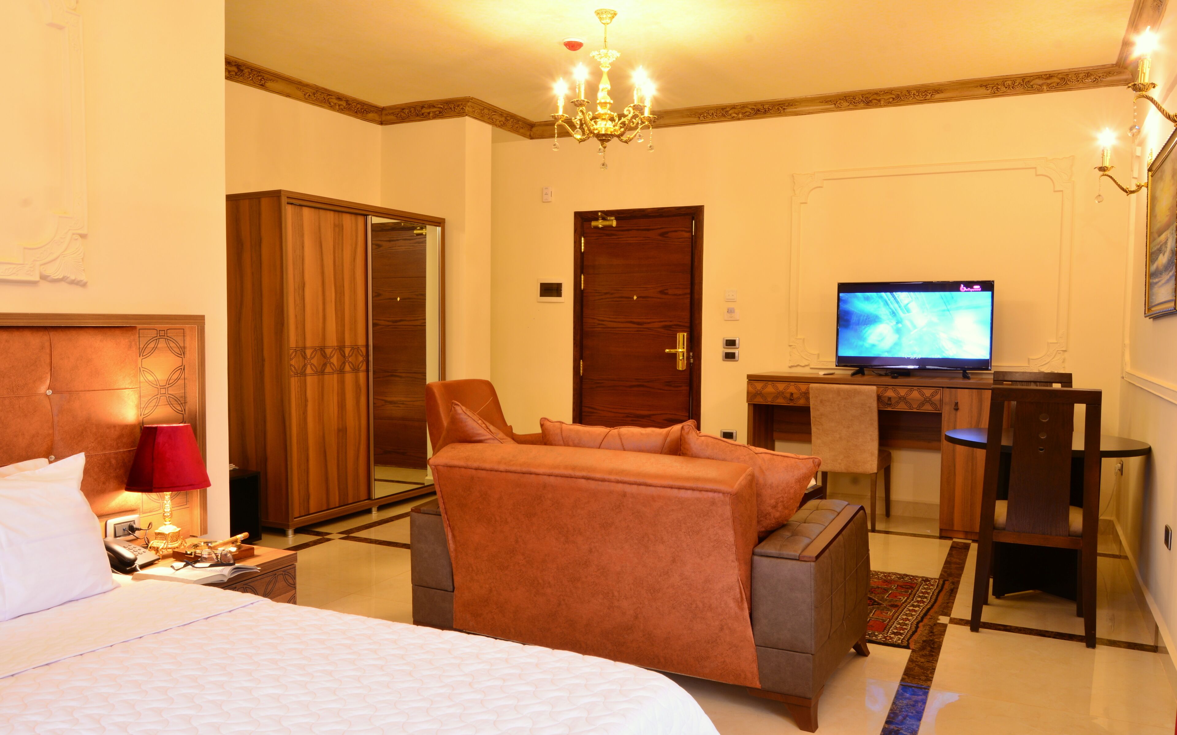 A hotel room with a bed, a sofa, a desk with a television, a wardrobe, and a chandelier.