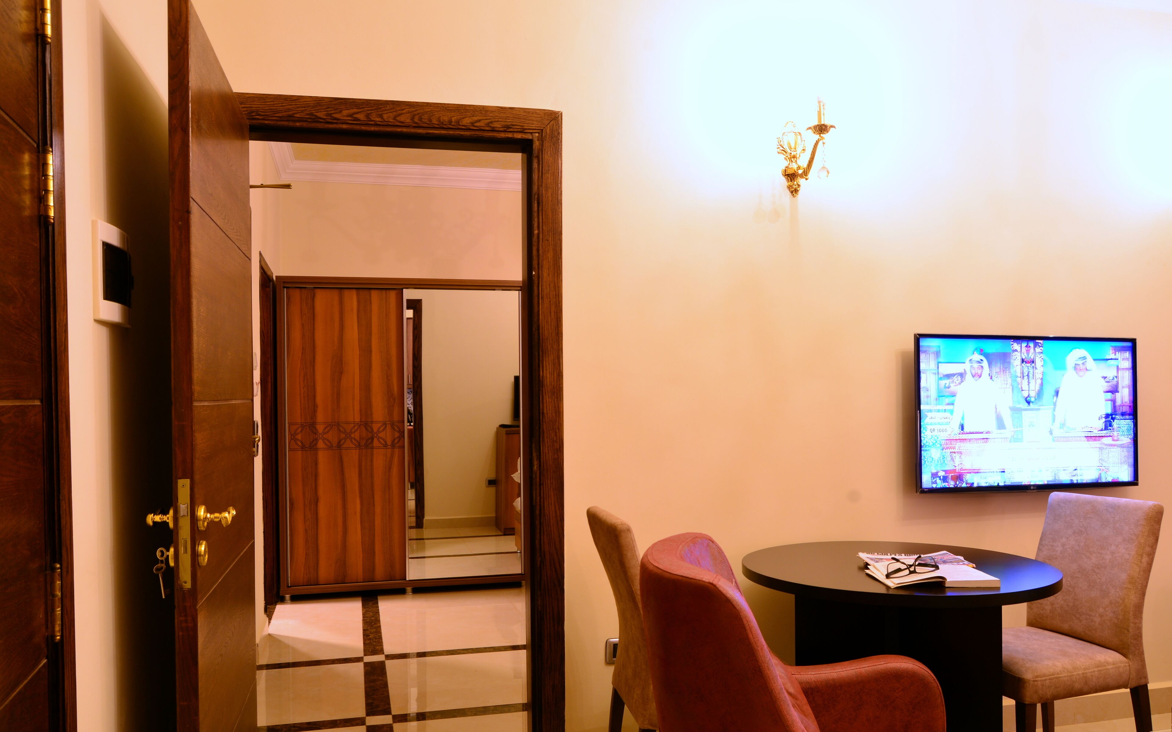 A hotel room with a wooden door, a checkered floor, a round table with two chairs, and a television on the wall.