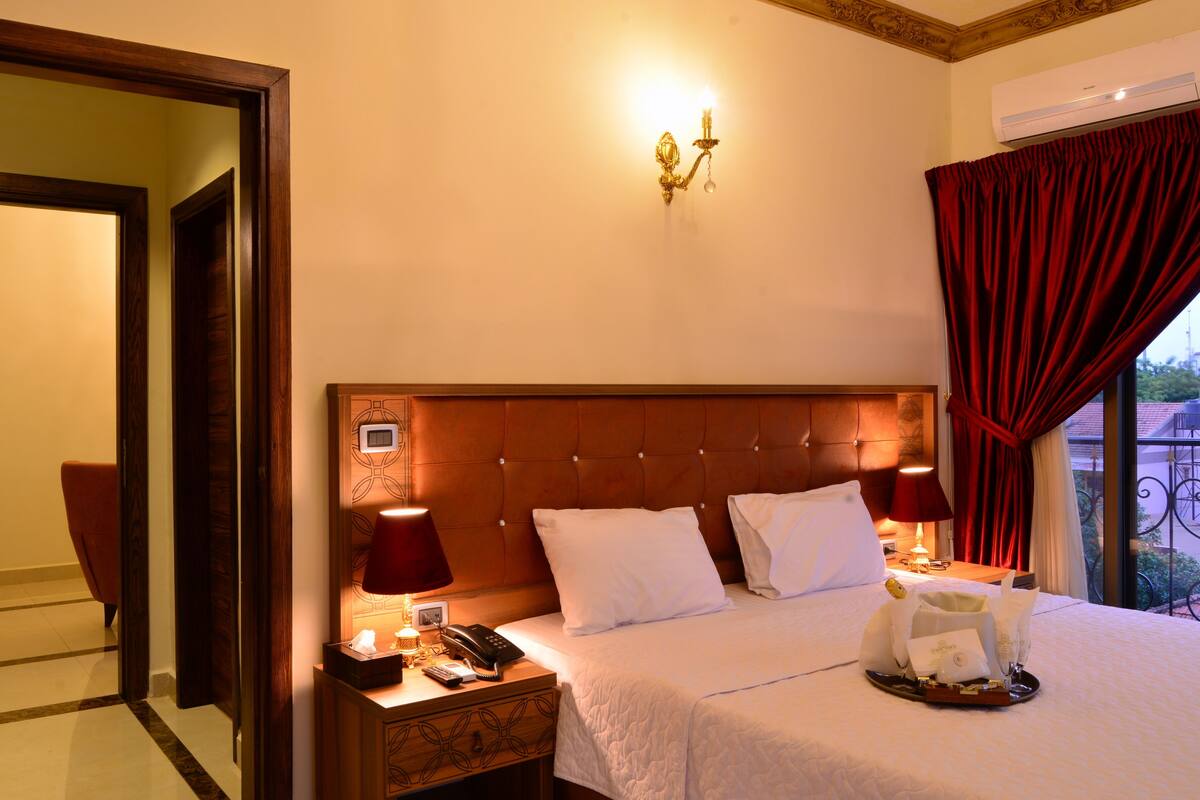 Hotelkamer met een groot bed, twee bedlampen, een nachtkastje met telefoon, een wandlamp en een raam met rode gordijnen.