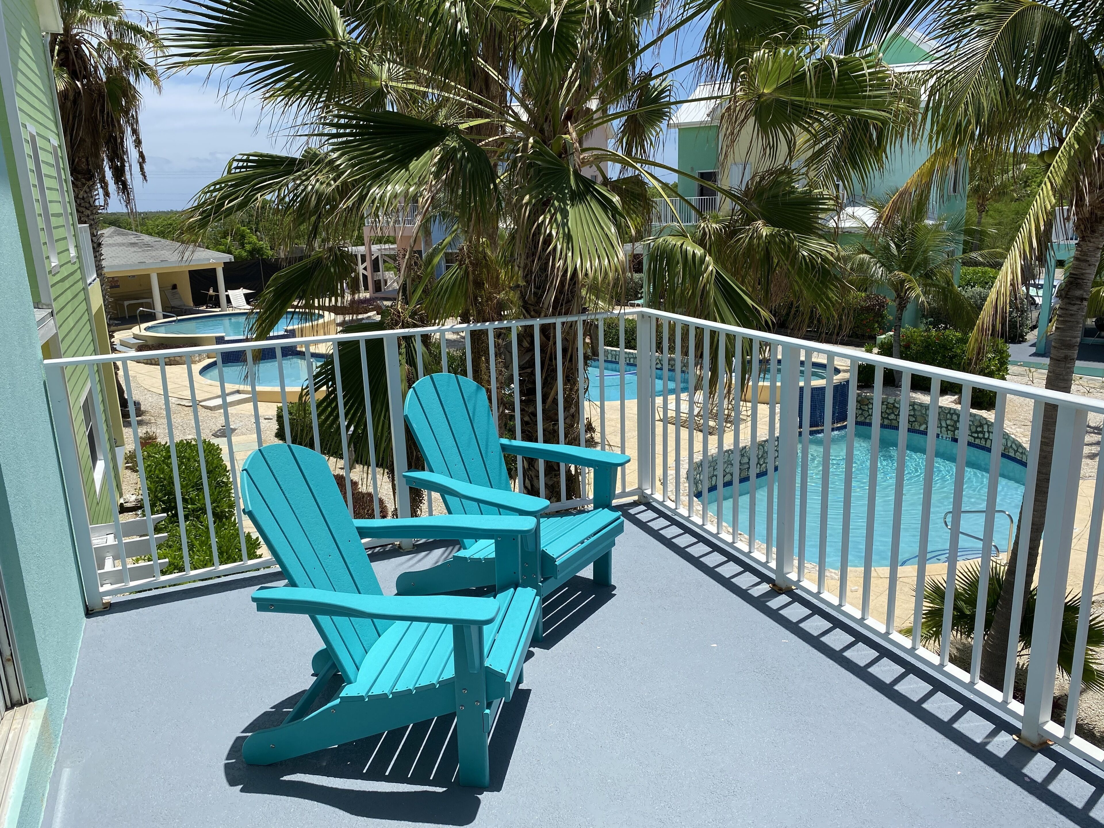 Un balcon avec des fauteuils Adirondack turquoise, une piscine et des palmiers.