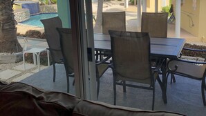 A view from inside a room with a glass door, looking out to a pool area with colorful buildings, palm trees, and a covered seating area.