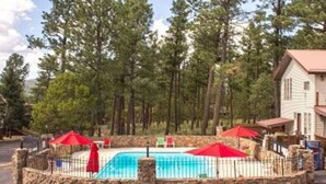 A pool area with a stone wall, red umbrellas, and a building with a red roof. - Raw Hide Lodge - Unit M1 (Ruidoso)