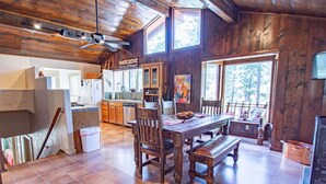 A rustic interior with a wooden dining table, chairs, and a bench. A kitchen area with cabinets and a refrigerator is visible in the background.