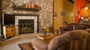 A cozy living room with a stone fireplace, comfortable seating, and a kitchen area visible in the background. - Come Stay in Elk Run - I2 (Ruidoso)
