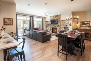 A spacious living area with a fireplace, a dining table set for four, and a large window overlooking a deck.