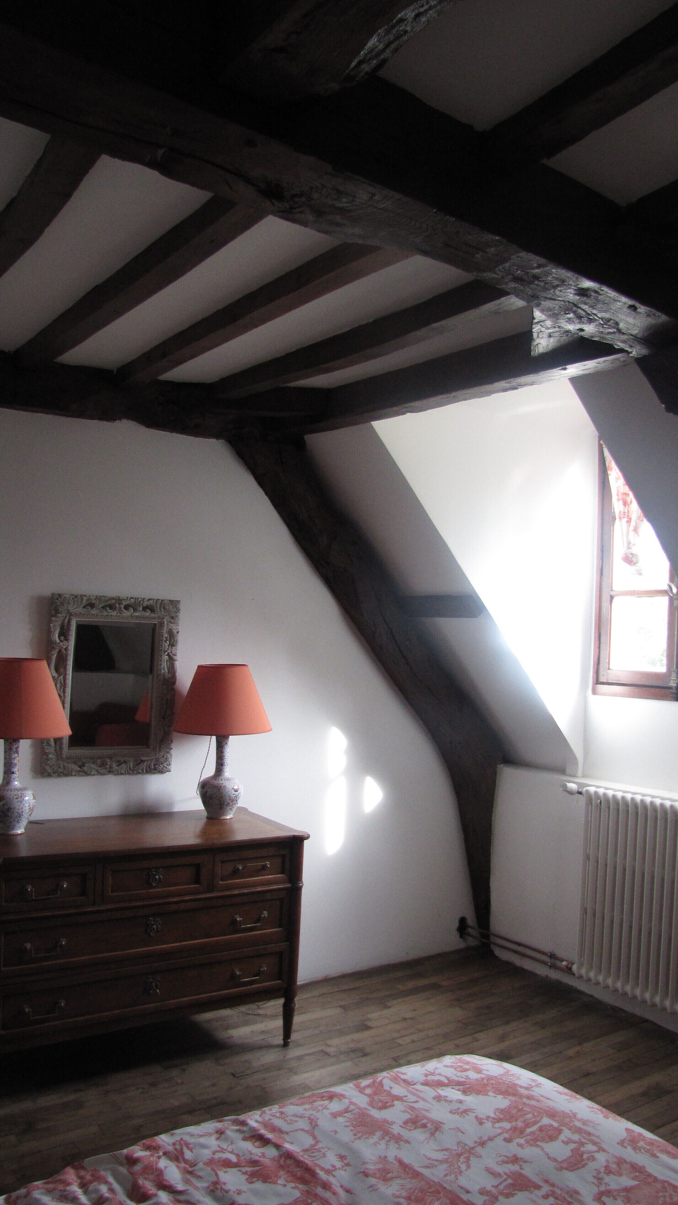 A room with a sloped ceiling, wooden beams, a dresser with lamps, a mirror, and a radiator.