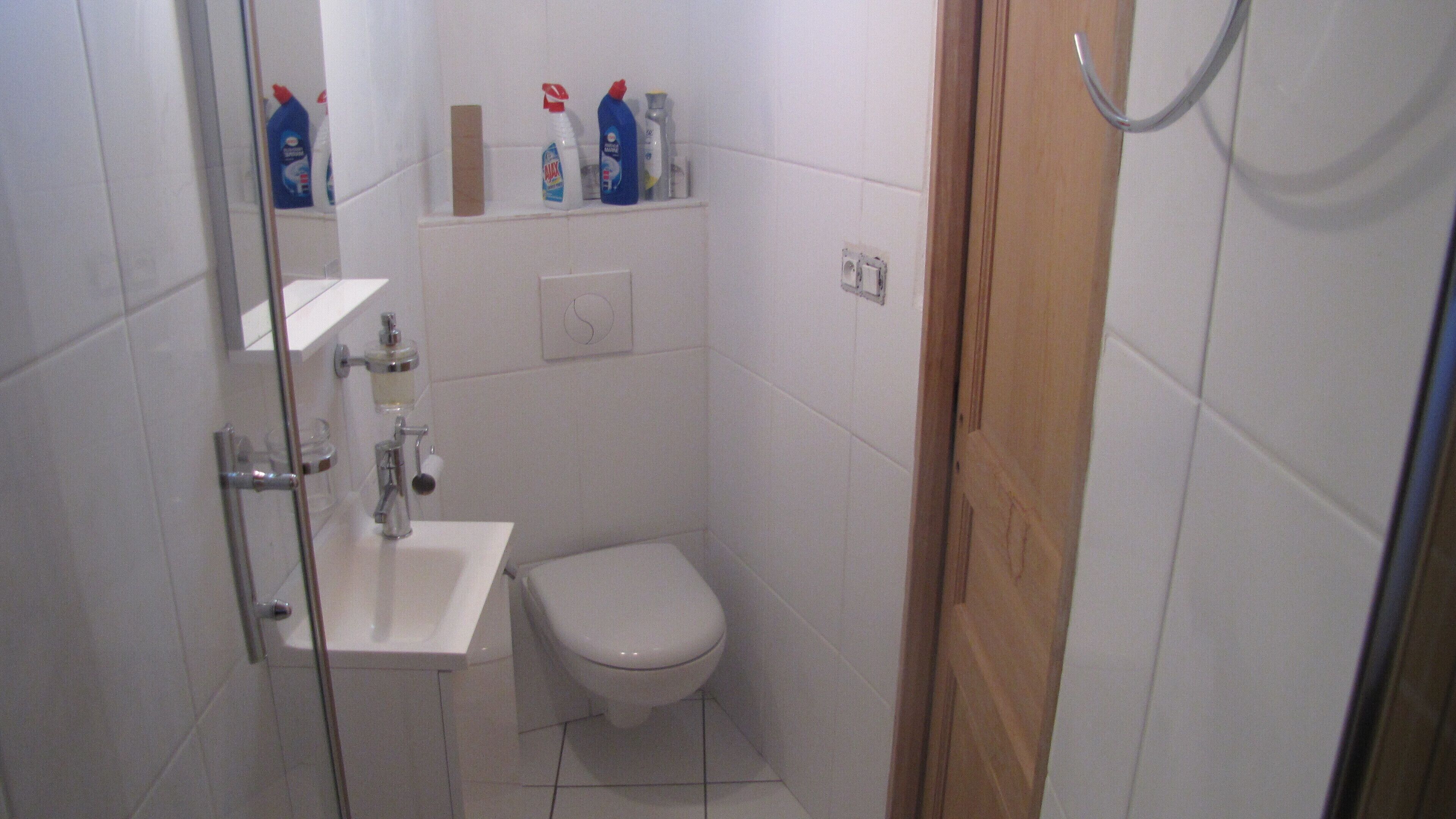 A small, white-tiled bathroom with a toilet, a sink, and a shower. There are toiletries on a shelf above the sink.