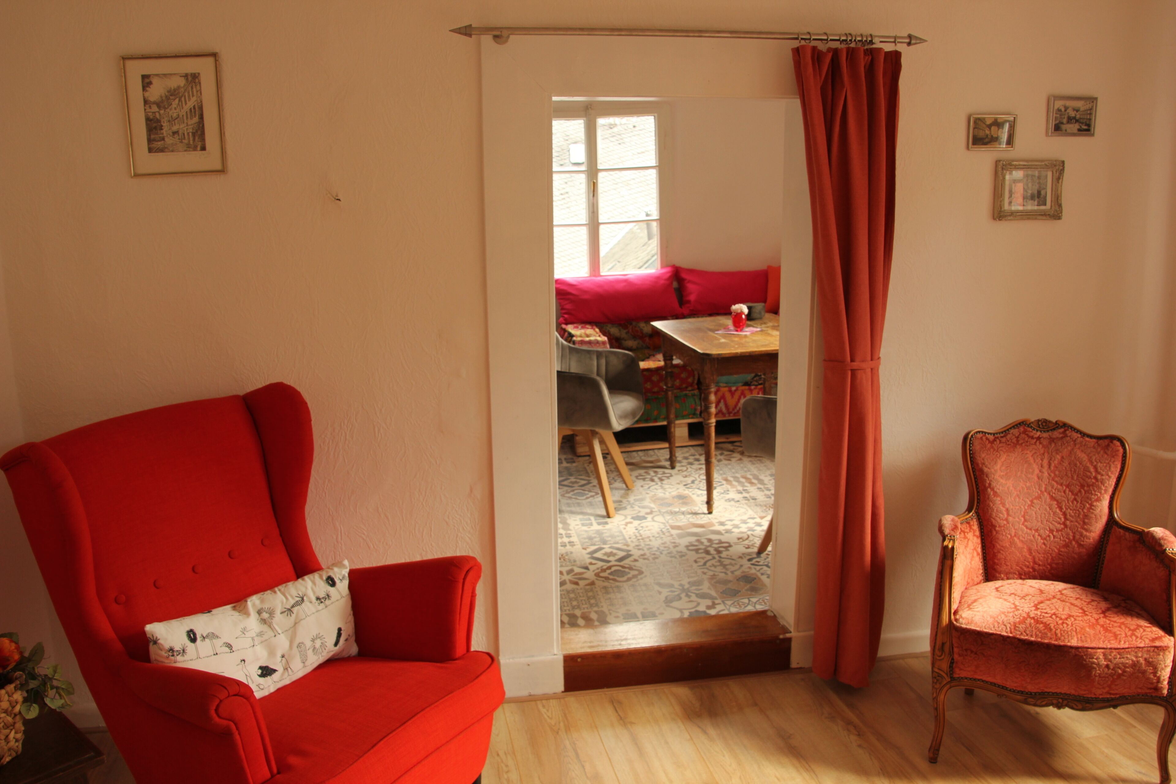 A room with a red armchair, a wooden floor, a door leading to a dining area, and a window with curtains.