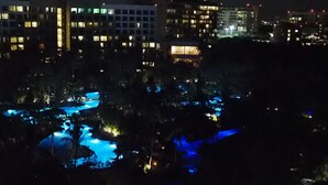 En nattbild av ett hotell med flera byggnader, ett poolområde med blå belysning och en avlägsen vy över en stadsbild.
