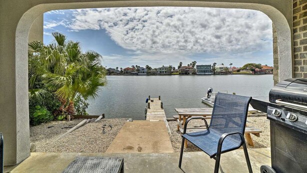 A patio with a grill, a chair, and a view of a lake with palm trees and houses in the background.